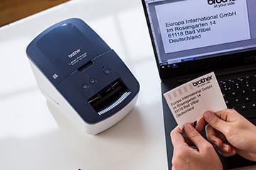 Printed address label with company logo being held in front of Brother QL-600B label printer