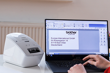 Brother QL-600R label printer next to a laptop with Brother P-touch Editor label design software on the screen, and label created ready for printing.