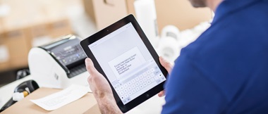 A man using Brother's iPrint&Label app on a tablet device to print a return shipping label on a Brother label printer, with packing boxes in the background.