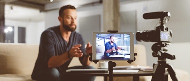 Male vlogger recording content on a digital camera with a tablet device displaying a preview while sitting on a sofa in front of a coffee table
