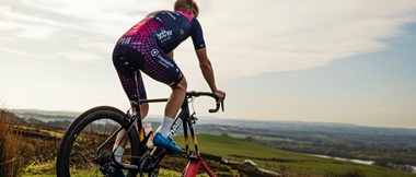 Cyclist on an outdoor trainer wearing a Crimson Performance jersey looking over fields with a cloudy sky in the background