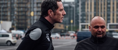 Side profile of Phil Jones MBE, stood next to Simon Howes on a city street with vehicles and tall buildings in the background