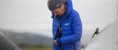 Cyclist Rebecca Richardson zipping up her blue coat while listening to music on earbuds with grey sky in the background
